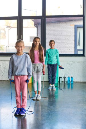Photo for Vertical shot of pretty little girls and boy with jump ropes in hands smiling at camera, child sport - Royalty Free Image