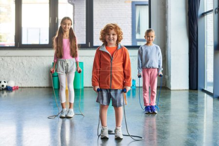 rothaariger Junge und zwei hübsche Mädchen posieren mit Springseilen und lächeln in die Kamera, Kindersport