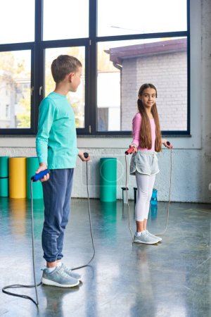 vertical shot of pretty children with skipping ropes in hands smiling at each other, child sport