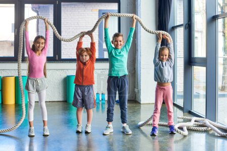 cheerful kids in sportswear raising fitness rope above heads and smiling at camera, child sport