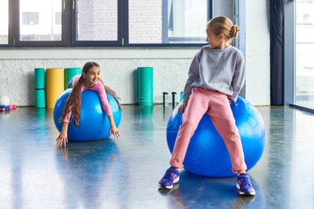 two cheerful small girls exercising on fitness balls and smiling at each other, child sport puzzle 677583450