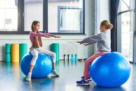 Foto de Dos niñas bonitas sentadas en pelotas de fitness delante de la otra con los brazos hacia adelante, deporte - Imagen libre de derechos