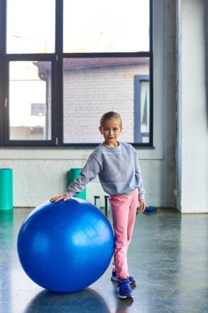 cute preadolescent girl in sportswear standing next to fitness ball and looking at camera, sport