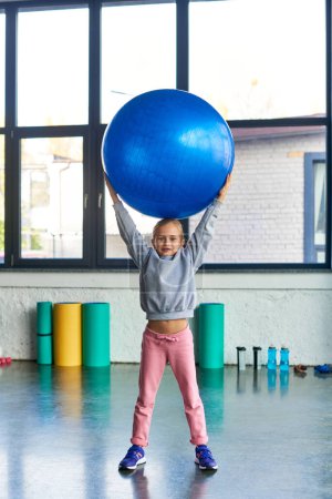 little cute girl in sportswear holding fitness ball above head and looking at camera, child sport