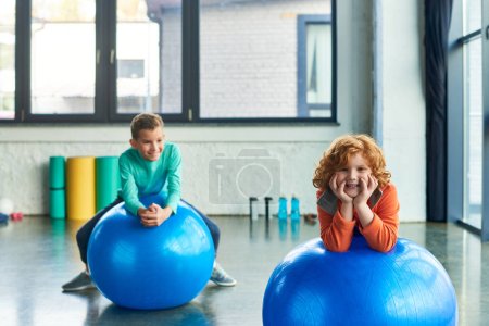 two joyful preadolescent boys exercising on fitness balls and smiling cheerfully, child sport