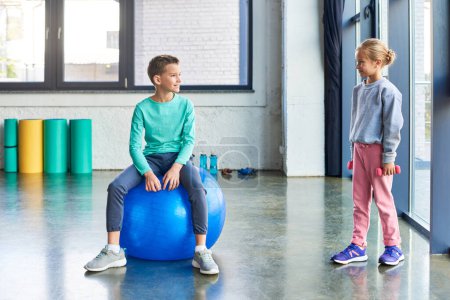 little blonde girl with dumbbells in hands smiling at cute boy on fitness ball, child sport