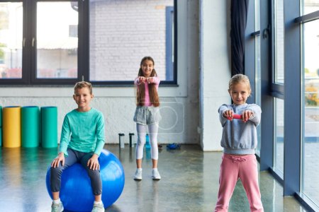 lindo niño sentado en la pelota de fitness con dos chicas bonitas haciendo ejercicio con pesas, deporte infantil