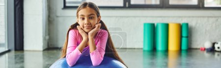 lustiges kleines Mädchen mit langen Haaren posiert mit Fitnessball mit Händen unterm Kinn, Kindersport, Banner