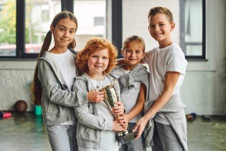vier fröhliche vorpubertäre Kinder in grauer Sportbekleidung posieren mit Trophäe in der Turnhalle, Kindersport
