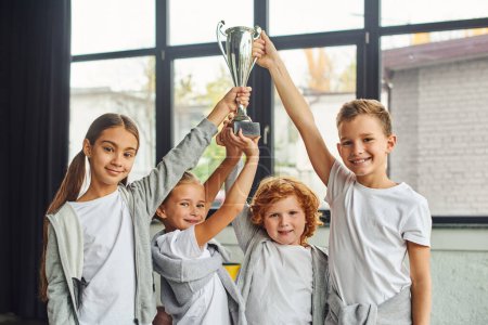 Photo for Cheering preadolescent boys and girls in sportswear raising trophy above heads, child sport - Royalty Free Image