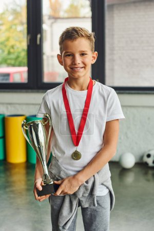 tiro vertical de niño lindo con medalla de oro y trofeo sonriendo a la cámara, deporte infantil