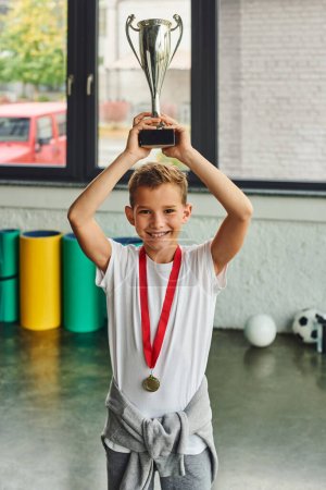 Photo for Vertical shot of cheerful little boy with golden medal raising trophy above his head, child sport - Royalty Free Image