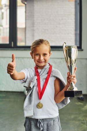 tiro vertical de chica rubia con trofeo y medalla de oro que muestra el pulgar hacia arriba en la cámara, deporte infantil