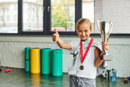 kleines blondes Mädchen posiert mit Trophäe und Goldmedaille und zeigt Daumen hoch in die Kamera, Kindersport