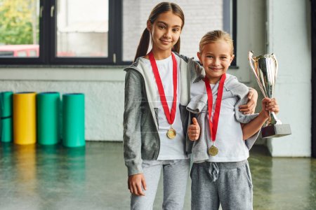 two cute preadolescent girls with medals holding trophy, smiling at camera, thumb up, child sport