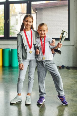vertical shot of cute preadolescent girls with medals and trophy hugging and smiling at camera
