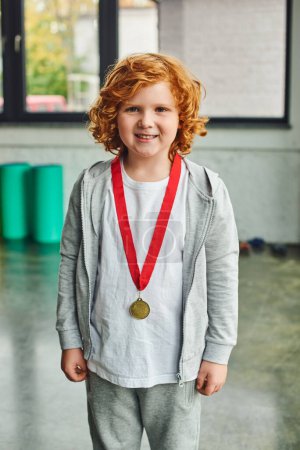 joyful red haired boy in sportswear with golden medal smiling sincerely at camera, child sport