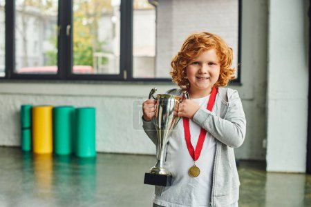 pretty red haired boy with golden medal holding huge trophy and smiling cheerfully at camera