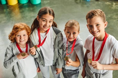 Foto de Cuatro niños lindos preadolescentes en ropa deportiva mostrando sus medallas de oro en la cámara, deporte infantil - Imagen libre de derechos