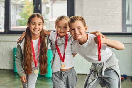 drei kleine Freunde in Sportbekleidung mit goldenen Medaillen, die sich umarmen und in die Kamera lächeln, Kindersport