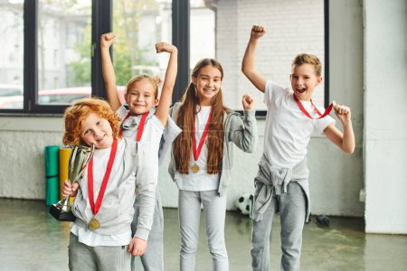 Photo for Cheering boys and girls in sportswear posing happily with trophy and golden medals, child sport - Royalty Free Image