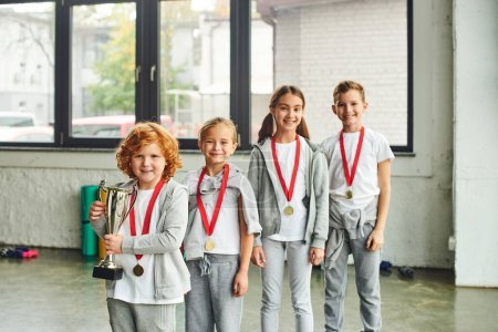 Photo for Cheerful little girls and boys smiling and posing with trophy and golden medal, child sport - Royalty Free Image
