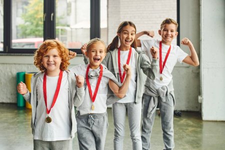 vier fröhliche Kinder in Sportkleidung mit Medaillen jubelnd und glücklich lächelnd in die Kamera, Kindersport