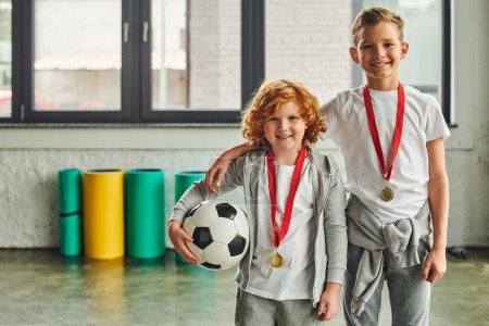 dos chicos alegres en ropa deportiva con medallas de oro sosteniendo pelota de fútbol y sonriendo a la cámara, deporte