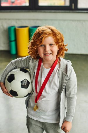 vertical shot of pretty red haired boy with golden medal holding soccer ball and smiling joyfully