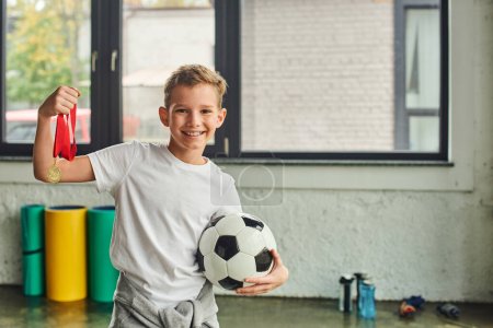 Photo for Cute cheerful preadolescent boy in sportswear holding golden medal and football, child sport - Royalty Free Image
