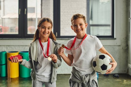 little girl with medal holding jump rope and cute boy posing with soccer ball, smiling at camera