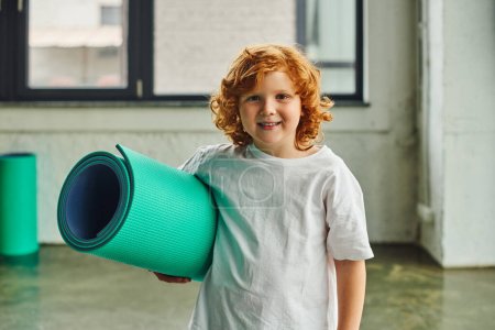 cute preadolescent boy with red hair posing with karemat in hands and smiling at camera, child sport
