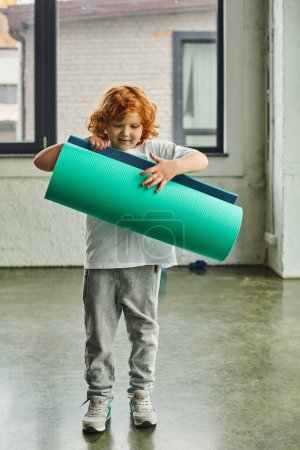 vertical shot of cute boy with red hair in sportswear unfolding his karemat, child sport