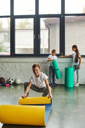 niedlichen vorpubertären fröhlichen Jungen entfaltet Fitnessmatte, während Mädchen auf Hintergrund stehen, Kindersport