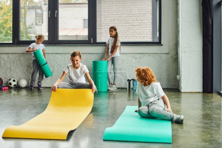 Photo for Cheerful preadolescent children in sportswear unfolding fitness mats in gym, workout, child sport - Royalty Free Image