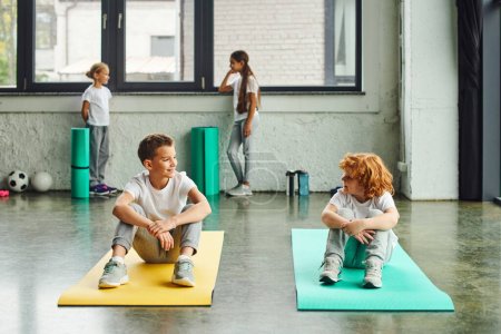 two cheerful boys smiling at each other on fitness mats with cute girls standing on backdrop