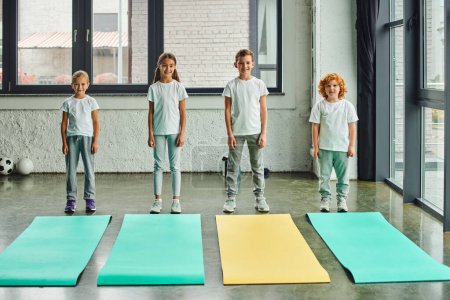 Photo for Jolly preadolescent boys and girls in sportswear posing next to fitness mats in gym, child sport - Royalty Free Image