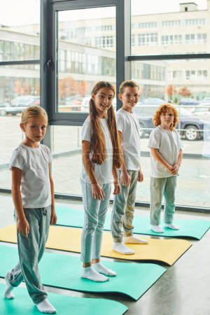 tiro vertical de niños alegres de pie sobre alfombras de fitness y sonriendo a la cámara, deporte infantil