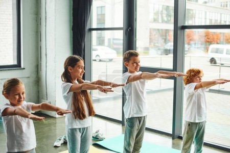 Photo for Joyous little children in sportswear stretching their arms in gym with window backdrop, child sport - Royalty Free Image