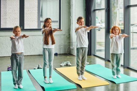 niños sonrientes felices estirando sus brazos de pie sobre alfombras de fitness en el gimnasio, deporte infantil