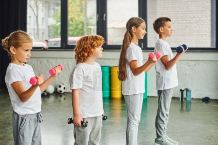 little children posing in profile holding dumbbells with fitness mats and balls on backdrop, sport