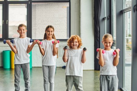 joyous preadolescent children exercising with dumbbells and smiling cheerfully at camera, sport