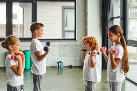 Photo for Pretty little children standing opposite each other and exercising with dumbbells, child sport - Royalty Free Image