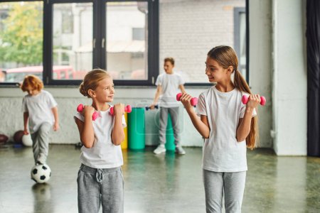 Photo for Little girls with dumbbells smiling at each other near blurred boys with soccer ball on backdrop - Royalty Free Image