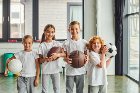 niños pequeños y alegres sosteniendo diferentes tipos de bolas y sonriendo a la cámara, deporte infantil