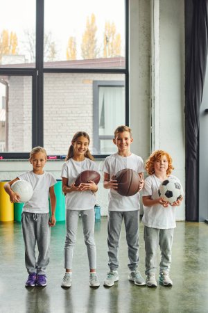 Photo for Vertical shot of four preadolescent children with different kinds of balls, smiling at camera, sport - Royalty Free Image