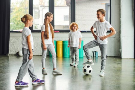 deux filles et un garçon roux souriant à un autre garçon mettant sa jambe sur le ballon de football, sport d'enfant