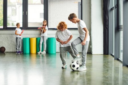 cheerful little boys playing football with girls holding dumbbells on background, child sport