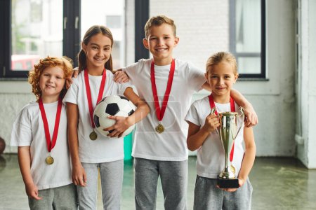 niños y niñas alegres con medallas sonriendo a la cámara y sosteniendo trofeo y fútbol, deporte