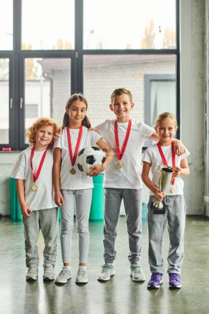 Senkrechte Aufnahme von vorpubertären niedlichen Kindern mit goldenen Medaillen, die mit Fußball und Trophäe posieren